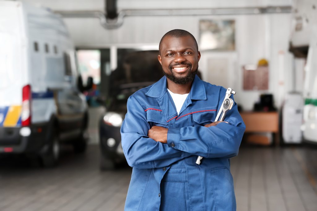 A smiling African-American automotive service professional in coveralls and holding two ratchets, standing in front of two vehicles in a repair bay. 