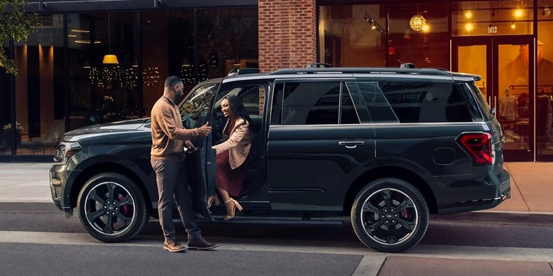 A man holding a door for a woman as she gets out of the driver's side of a black 2024 Ford Expedition in front of a hotel.