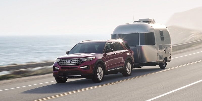 A red 2024 Ford Explorer towing a camping trailer down the highway backed by the ocean.