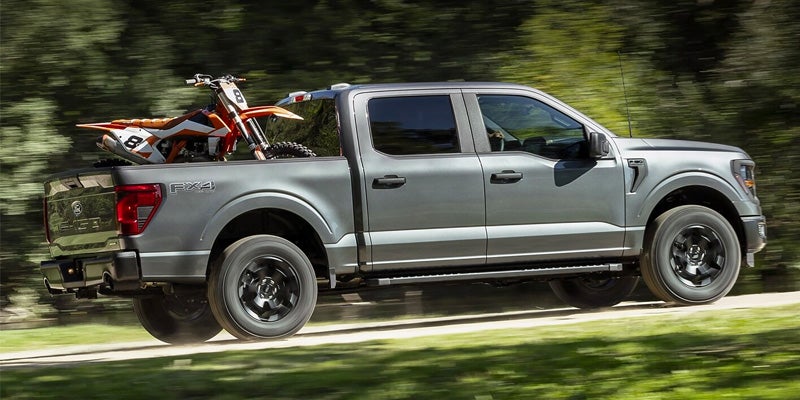 A green Ford F-150 driving down the road with a dirt bike in the bed.